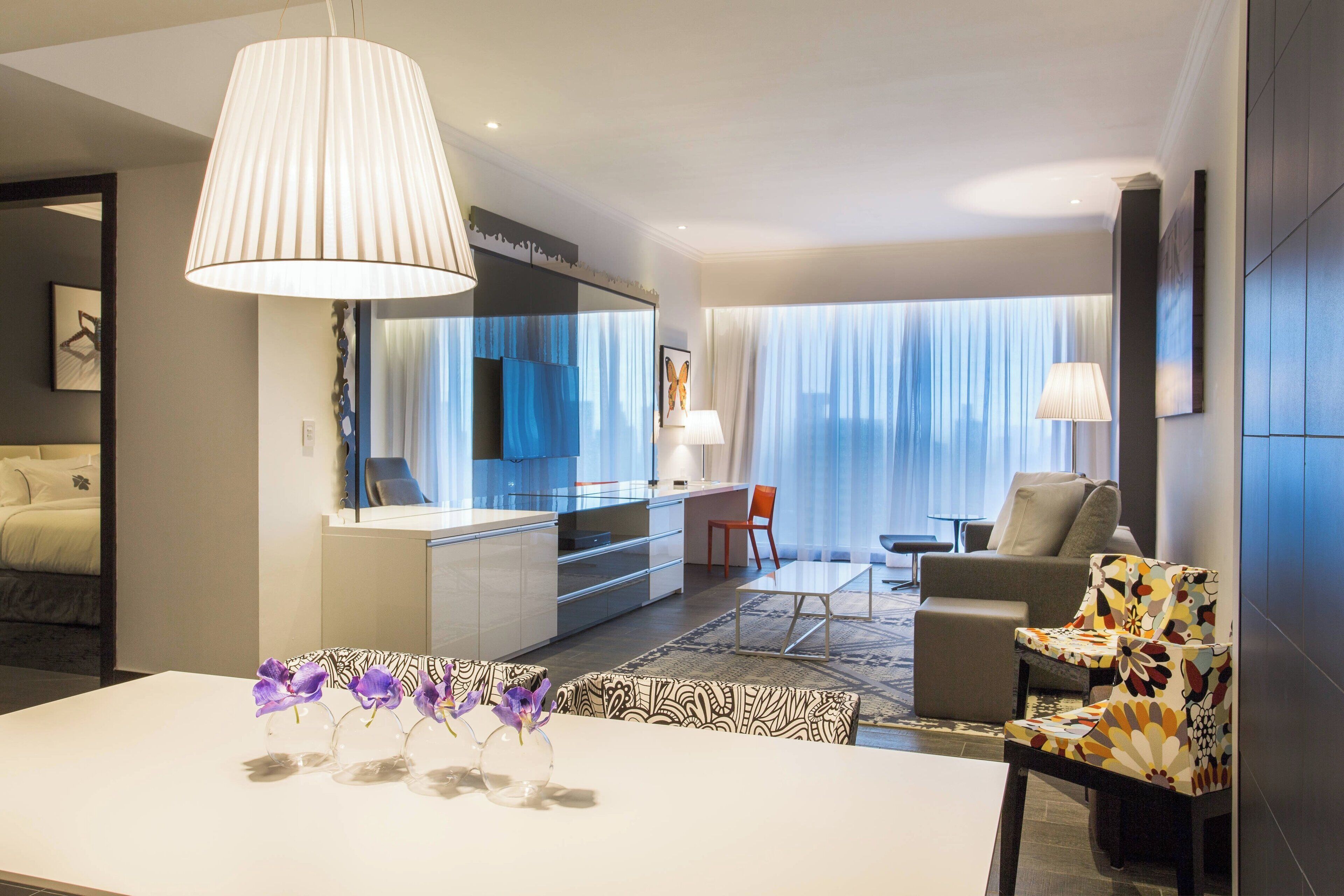 A modern hotel room with a dining area, a sofa, a chair, and a large window with curtains.