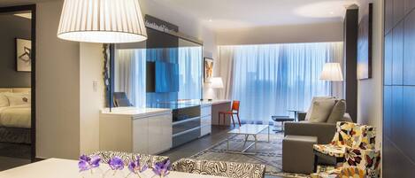 A modern hotel room with a dining area, a sofa, a chair, and a large window with curtains.
