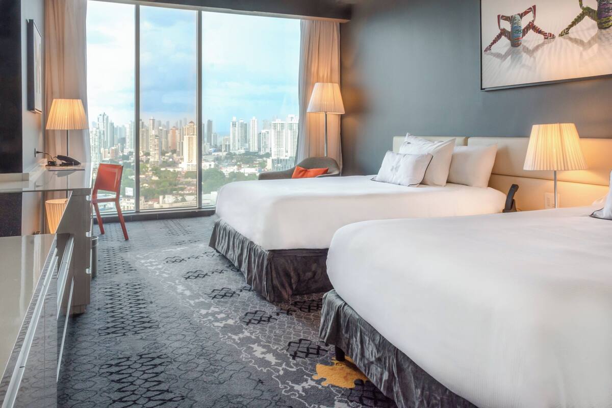 A hotel room with two beds, a desk, a chair, and a large window offering a city view.