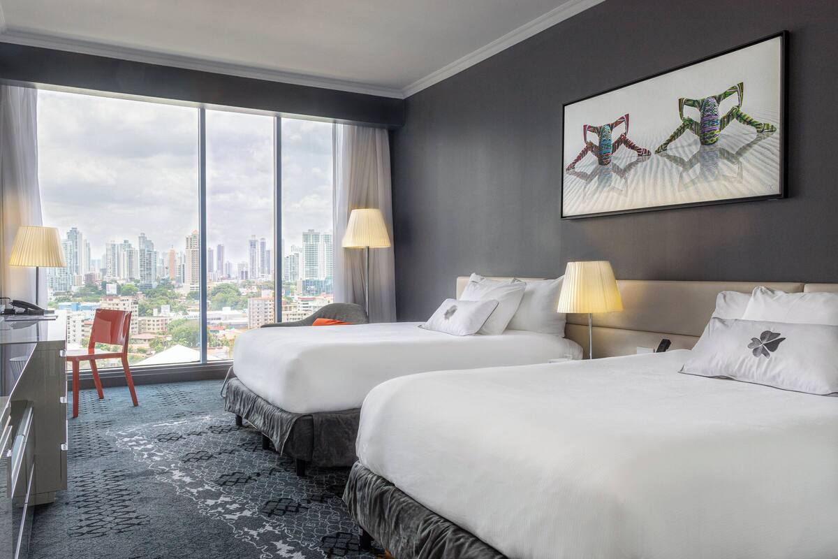 A hotel room with two beds, a desk, a chair, and a large window offering a city view.