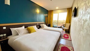 A hotel room with two beds, a chair, a TV, and a balcony with yellow curtains. - Hôtel Belle Vue & Spa (Meknes)