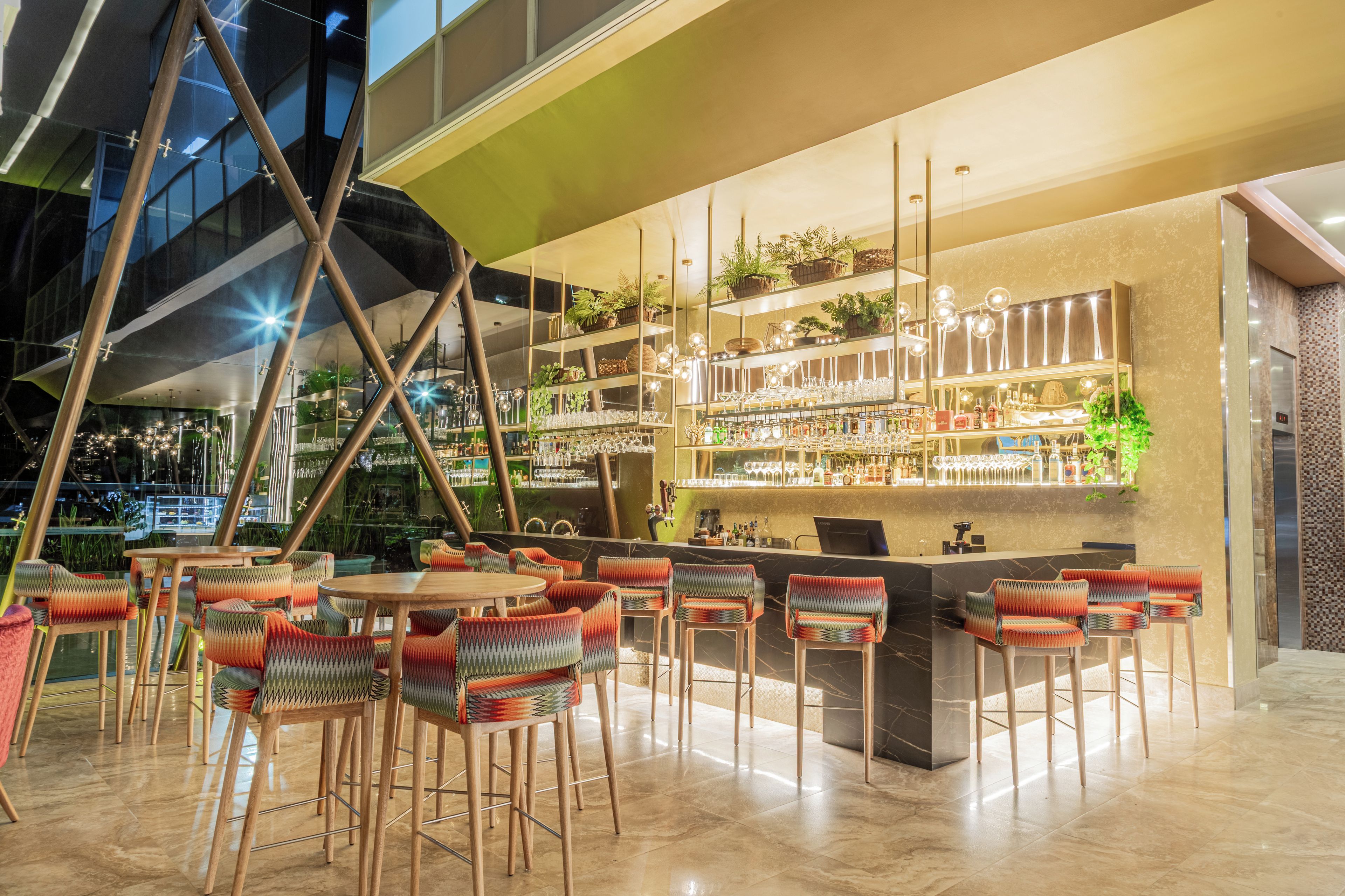 Zona de bar moderna con techo alto, taburetes y estantes llenos de botellas y vasos.