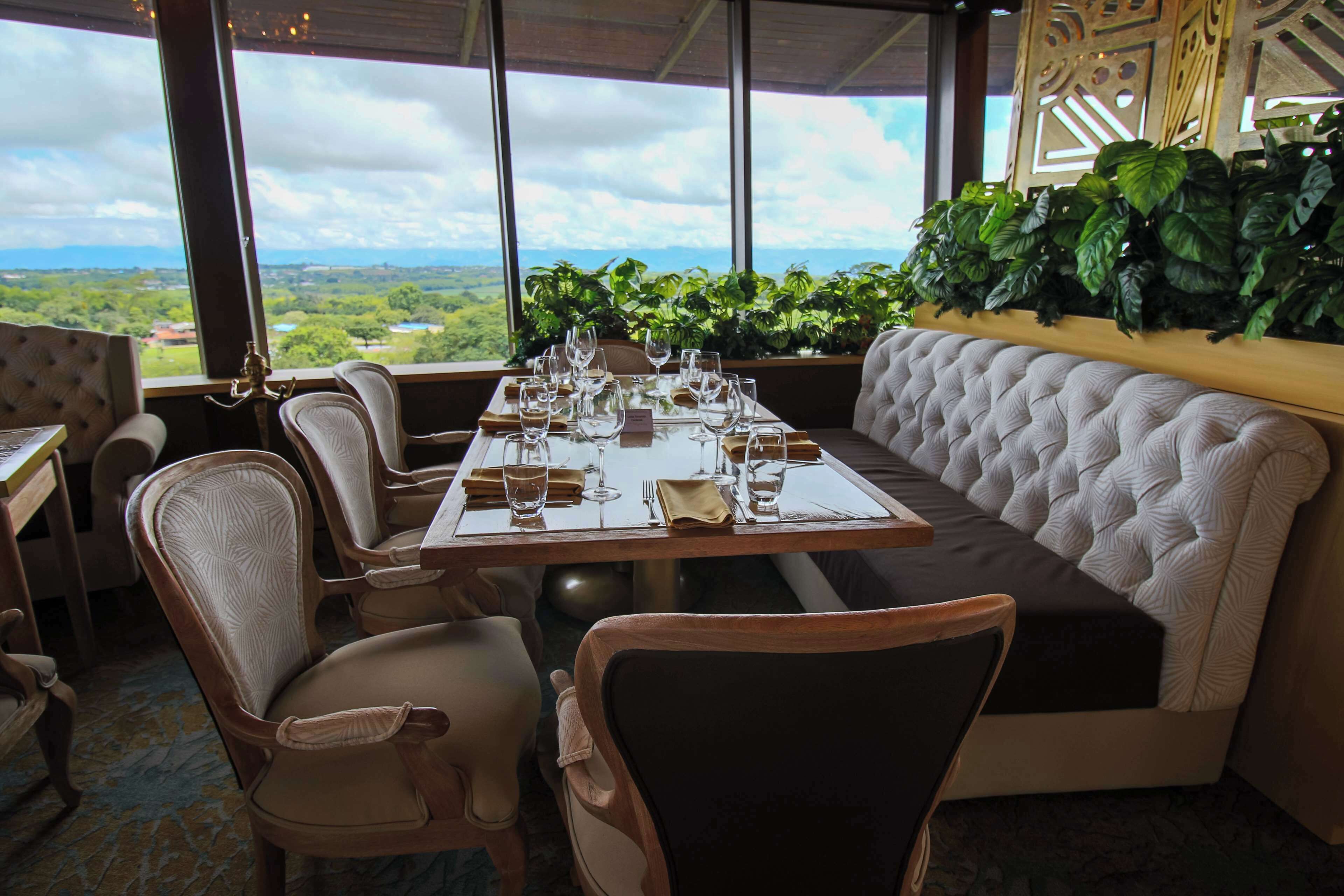 A dining area with a table set for four, surrounded by upholstered chairs, with a view of greenery and distant landscapes through large windows.