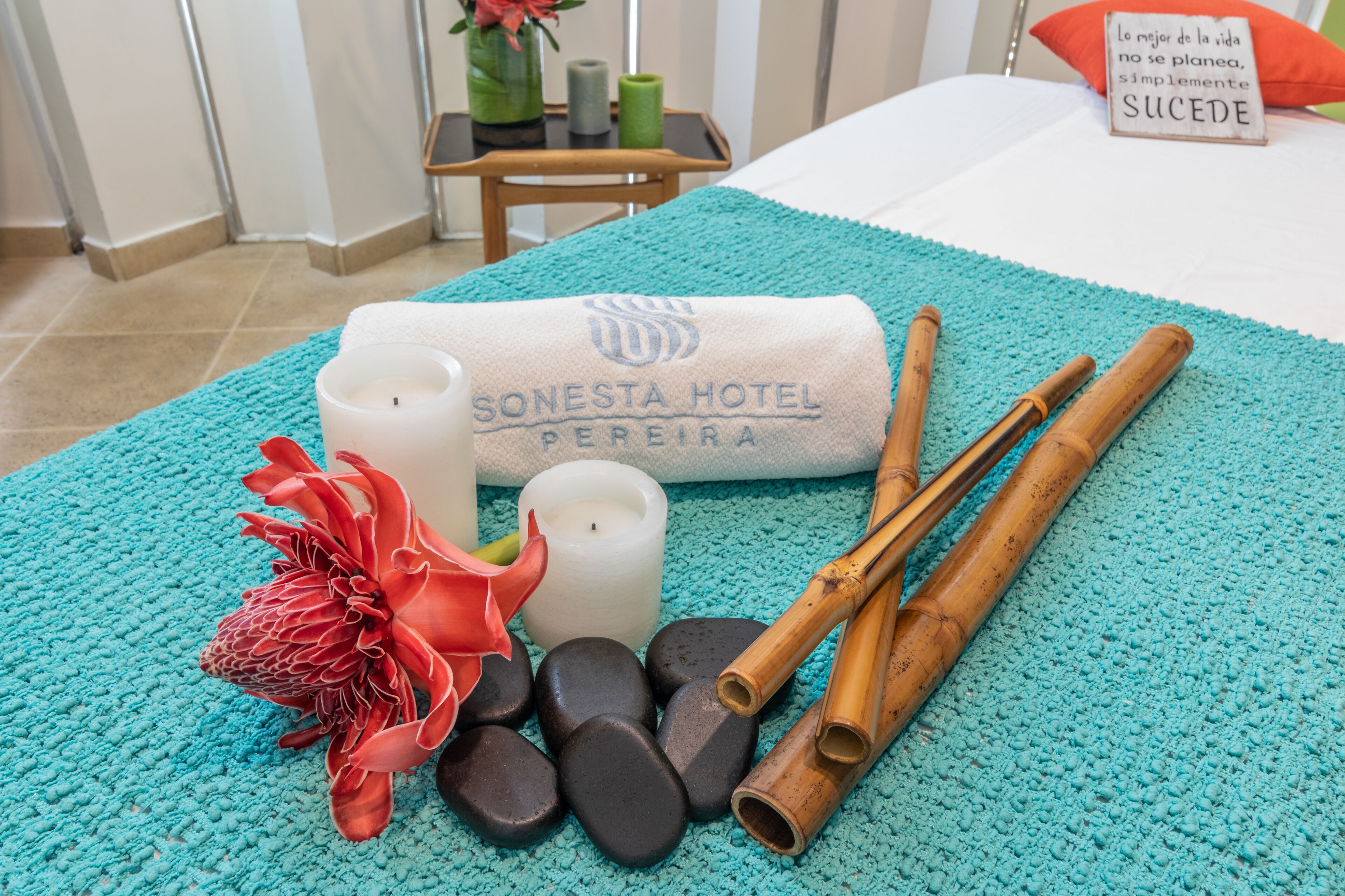 A room with a bed, a teal bath mat, a rolled towel, candles, a flower, and wooden sticks.