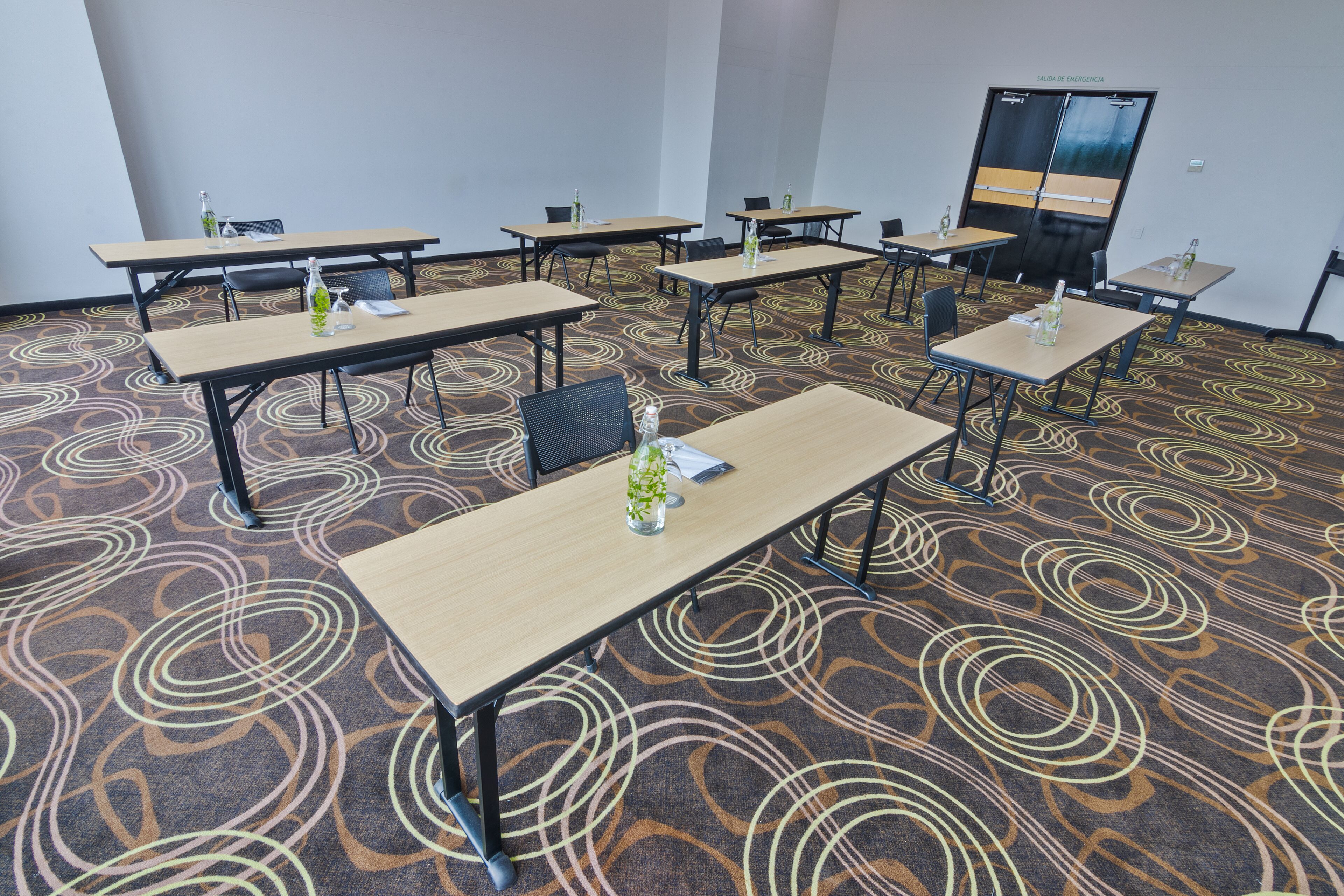 A conference room with multiple tables and chairs, a patterned carpet, and a door labeled "Sala de Emergencia".