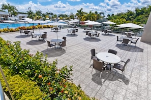 Relax and socialize in a spacious rooftop area with tables, chairs, and umbrellas, surrounded by lush plants and a scenic pool view.