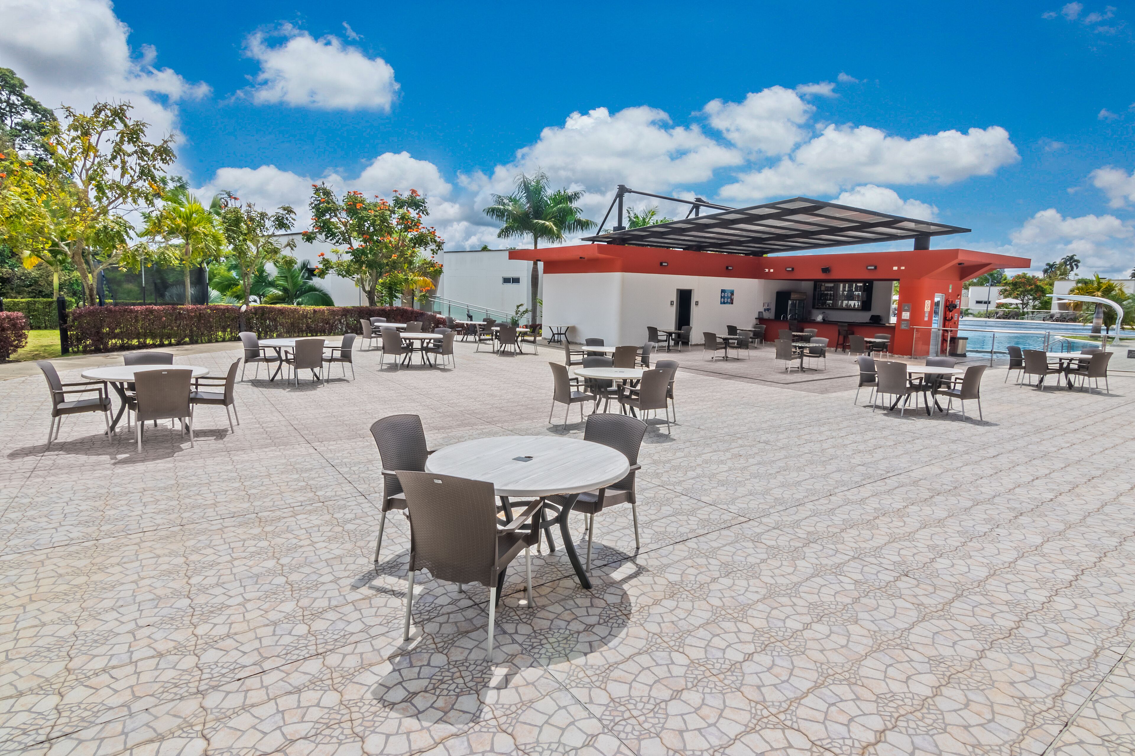 An outdoor patio area with multiple round tables and chairs, a bar area, and a pool in the background.