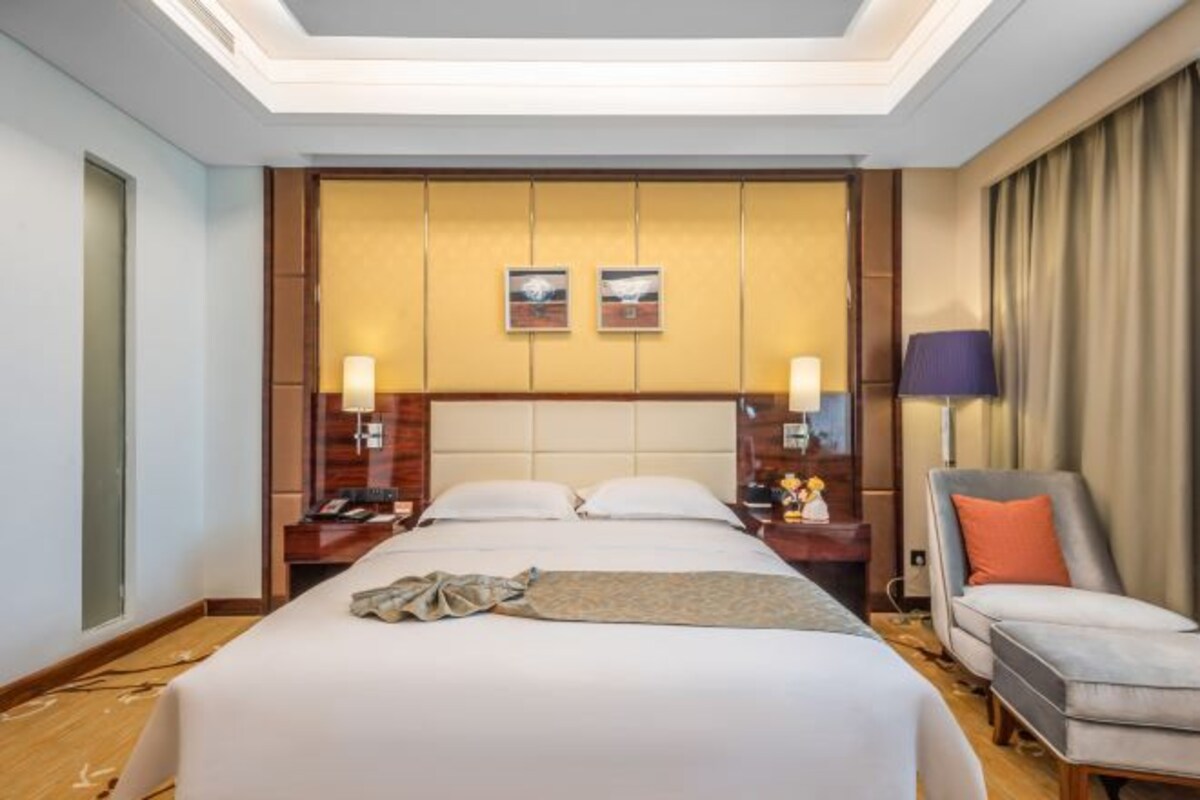 A hotel room with a large bed, two bedside tables, a chair, and a headboard with artwork.