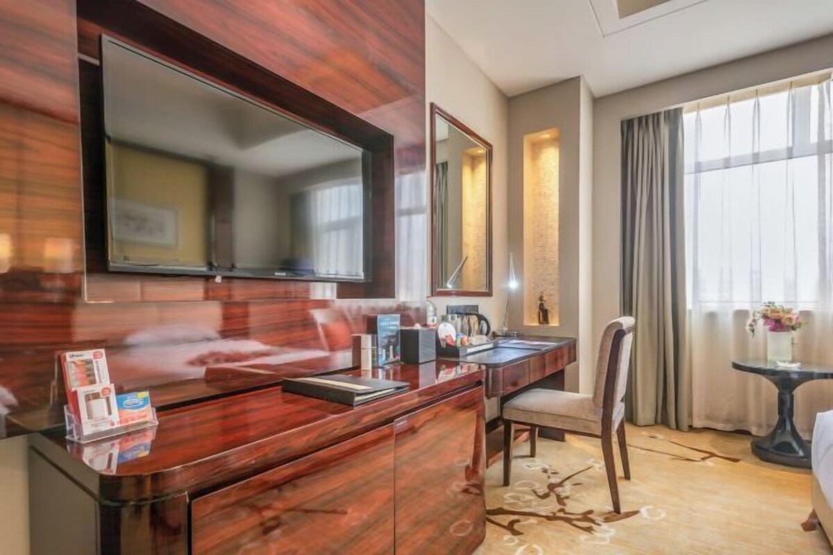 A hotel room with a wooden desk, a flat-screen TV, a mirror, a chair, a small table with a vase, and a large window with curtains.