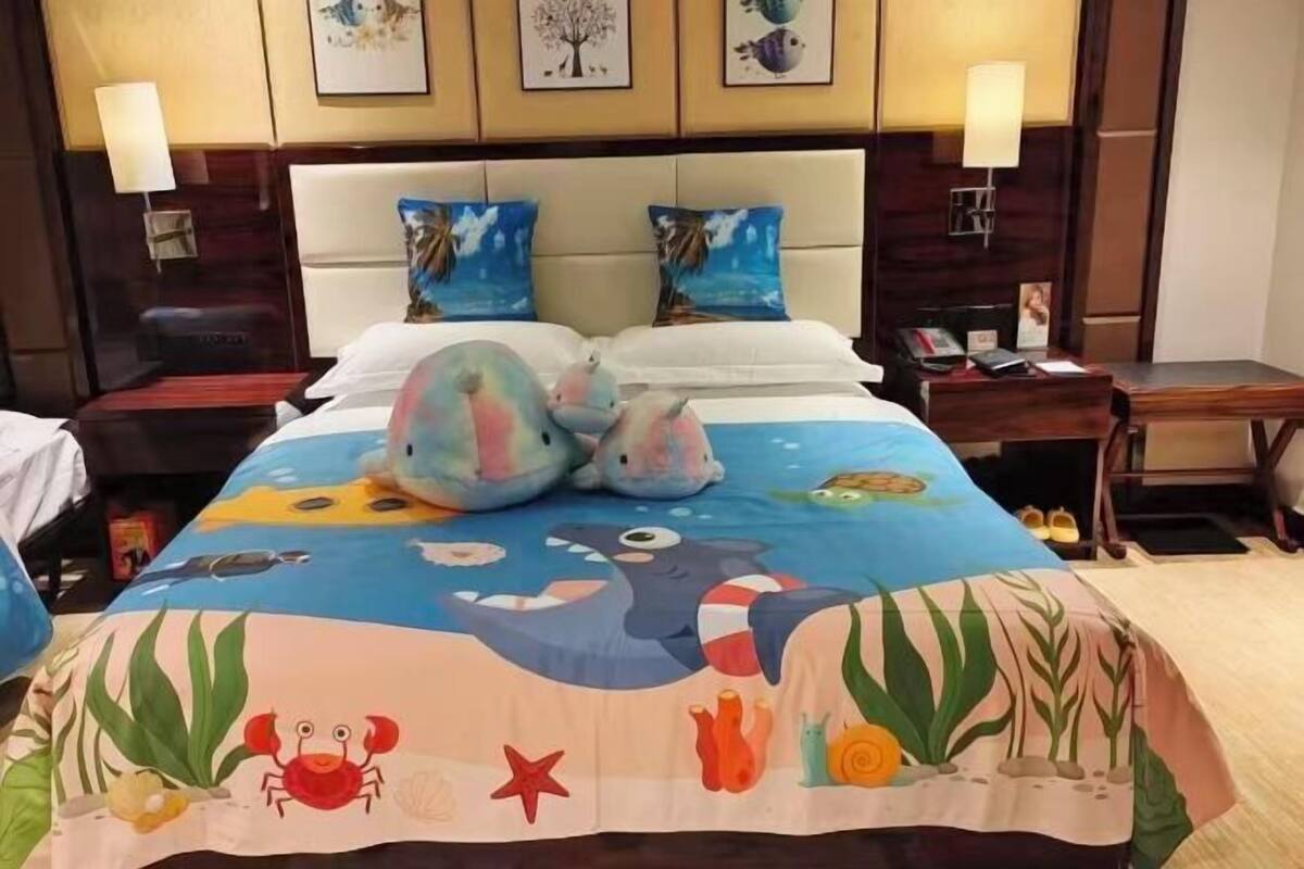 A hotel room with a bed featuring a colorful ocean-themed bedspread, a headboard with a light wood finish, and two bedside lamps.