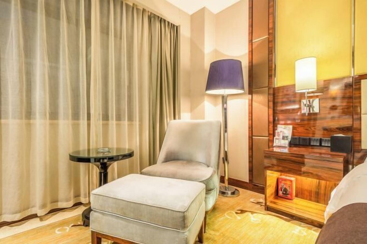 A hotel room with a bed, a chair, a small table, a lamp, a mirror, and a wooden dresser.