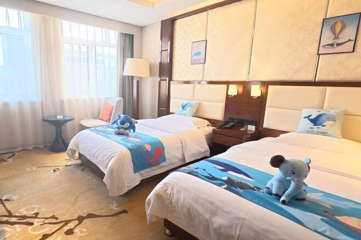 A hotel room with two beds, each with a plush toy and a blue-themed bedspread. There is a nightstand with a lamp, a window with curtains, and a wall with a framed picture.