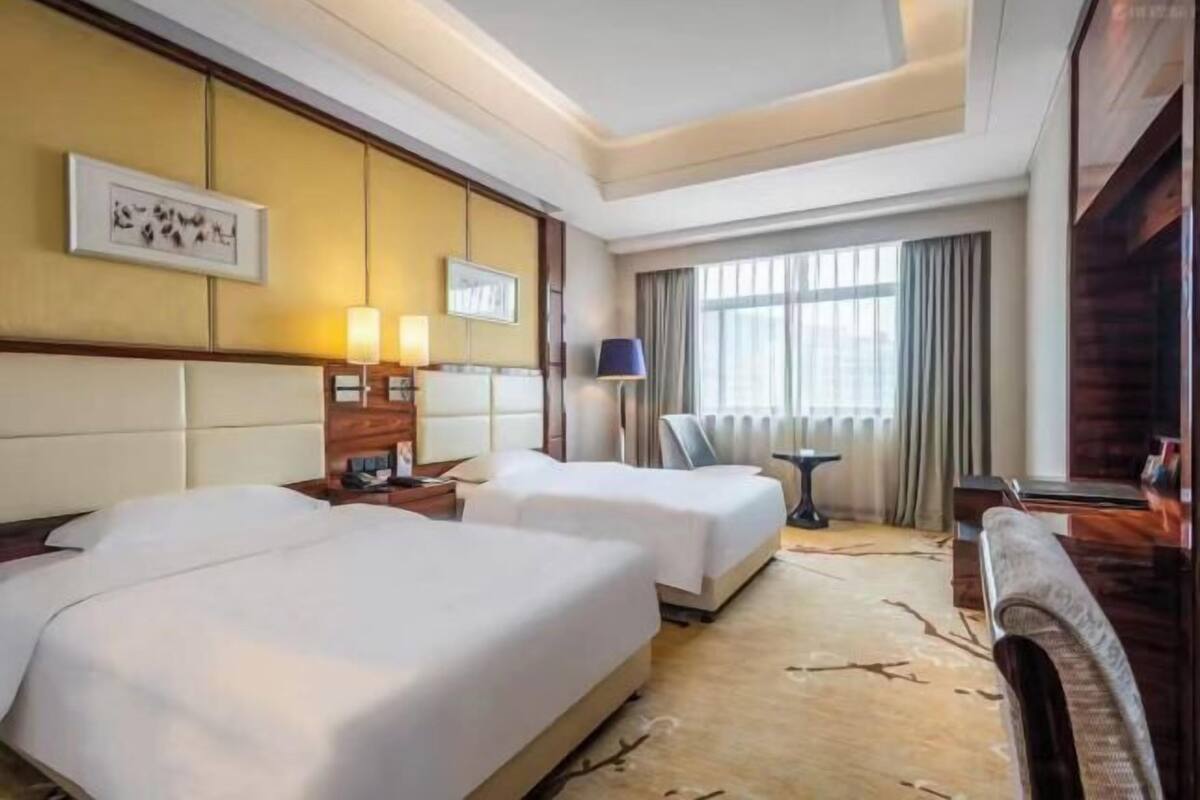 A hotel room with two beds, a small table, a chair, a lamp, and a large window with curtains.