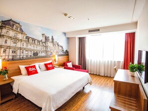 Habitación de hotel con una cama grande, almohadas rojas y blancas, cabecera de madera, un escritorio con una planta y un mural de un paisaje urbano en la pared.