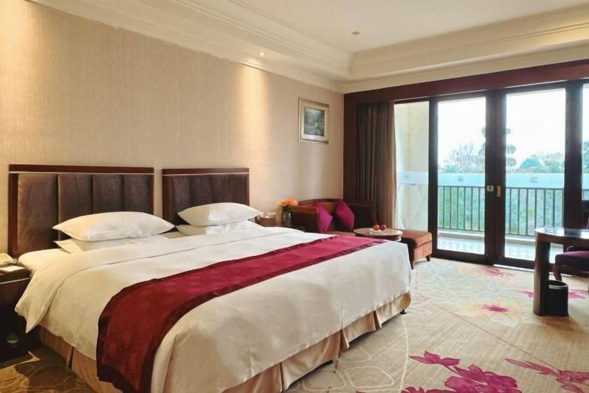 A hotel room with a large bed, a desk, a chair, a balcony with a view, and a door.