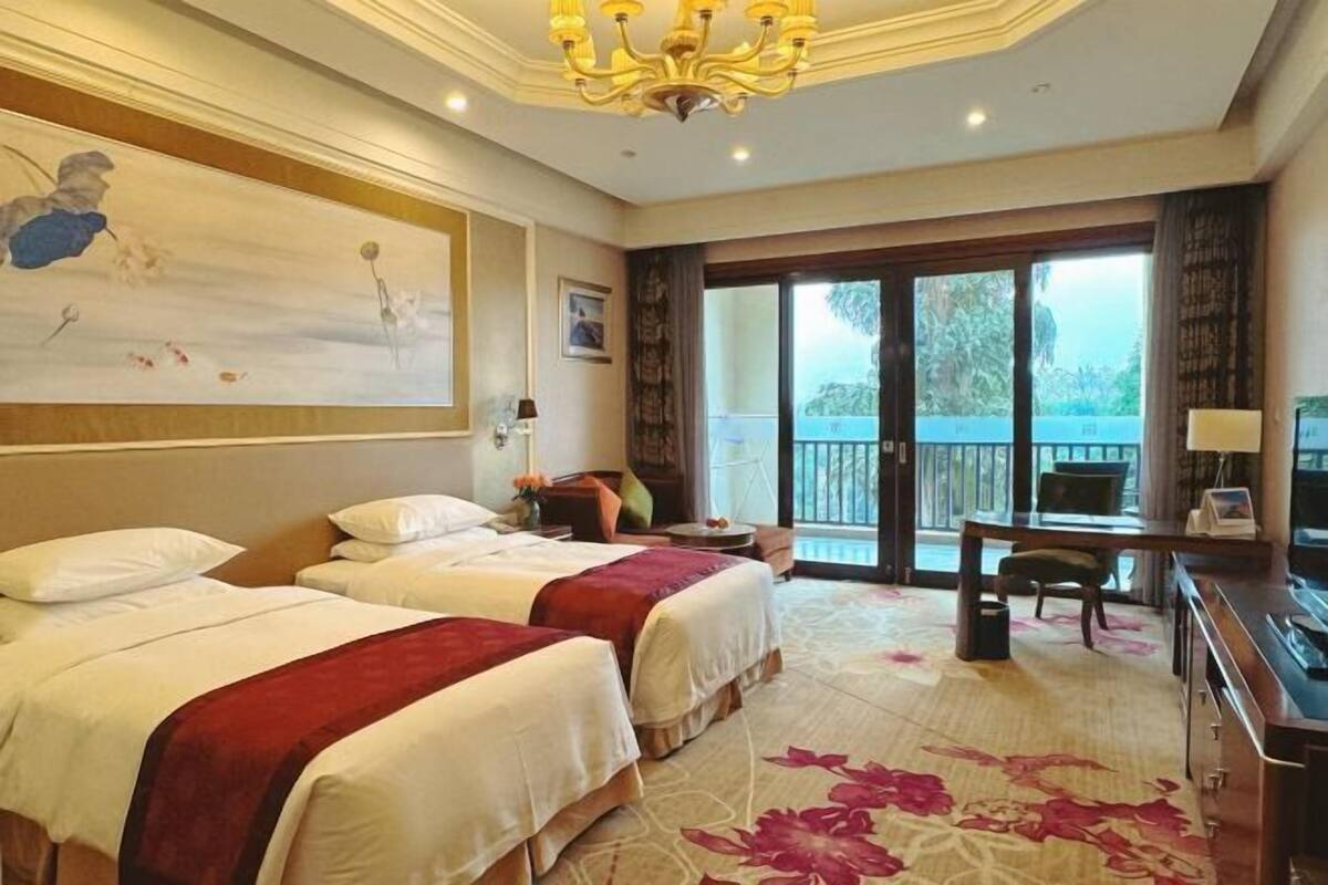 A hotel room with two beds, a desk, a chair, a balcony with a view, and a chandelier.
