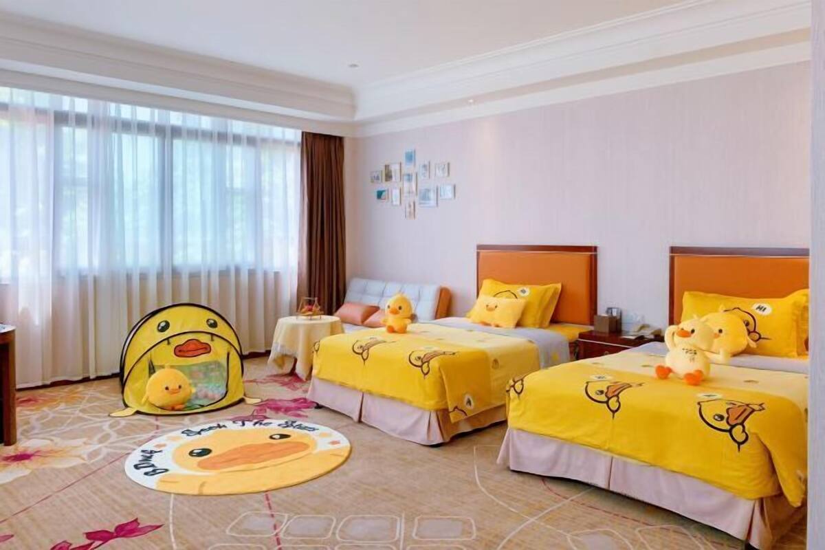 A hotel room with two beds, each with a yellow duck-themed bedspread and pillows. There is a round rug with a duck design on the floor, and a window with curtains.