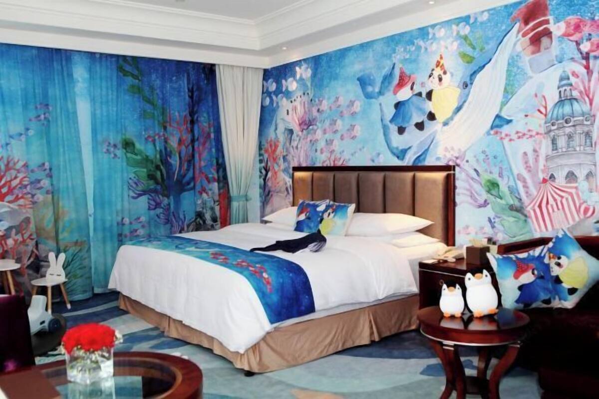 A hotel room with a large bed, a nightstand, a round table with two penguin-shaped lamps, and a mural on the wall.