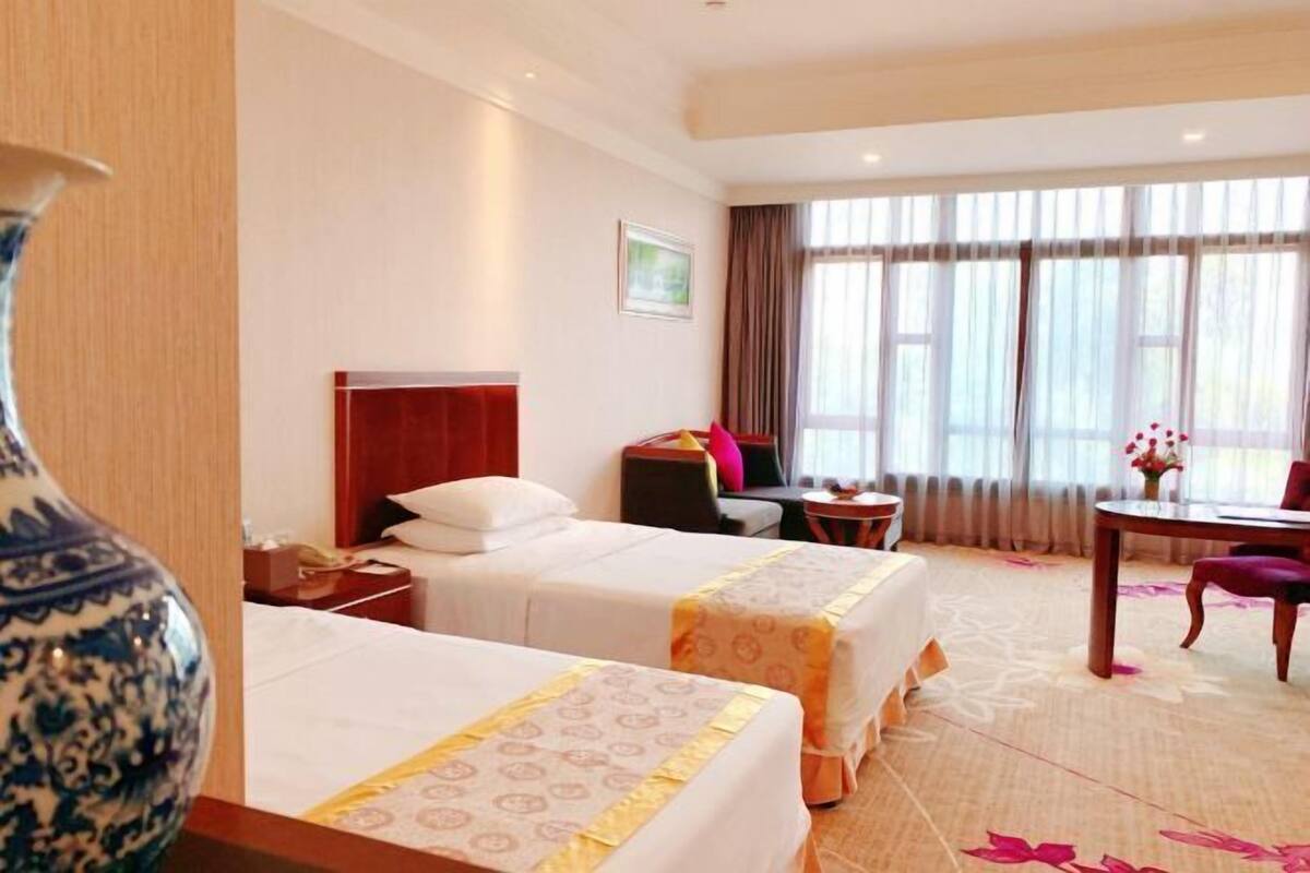 A hotel room with two beds, a desk, a chair, a vase, and a window with curtains.