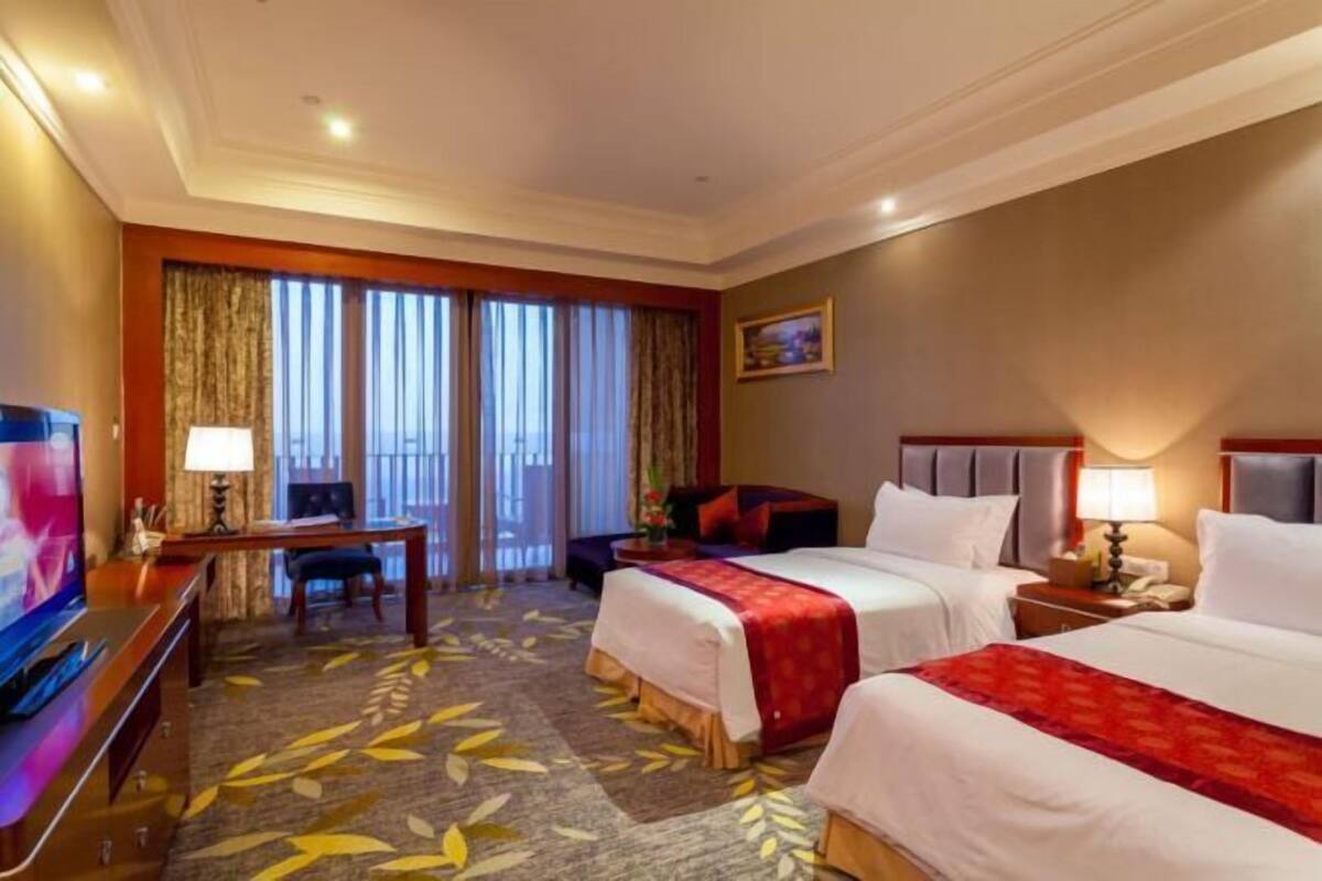 A hotel room with two beds, a desk, a TV, and a balcony with curtains.