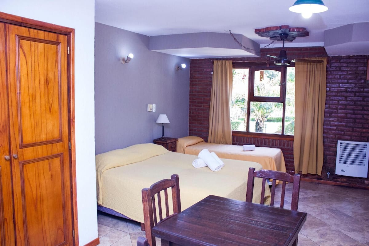 A hotel room with two beds, a wooden table, a ceiling fan, and a window with curtains.