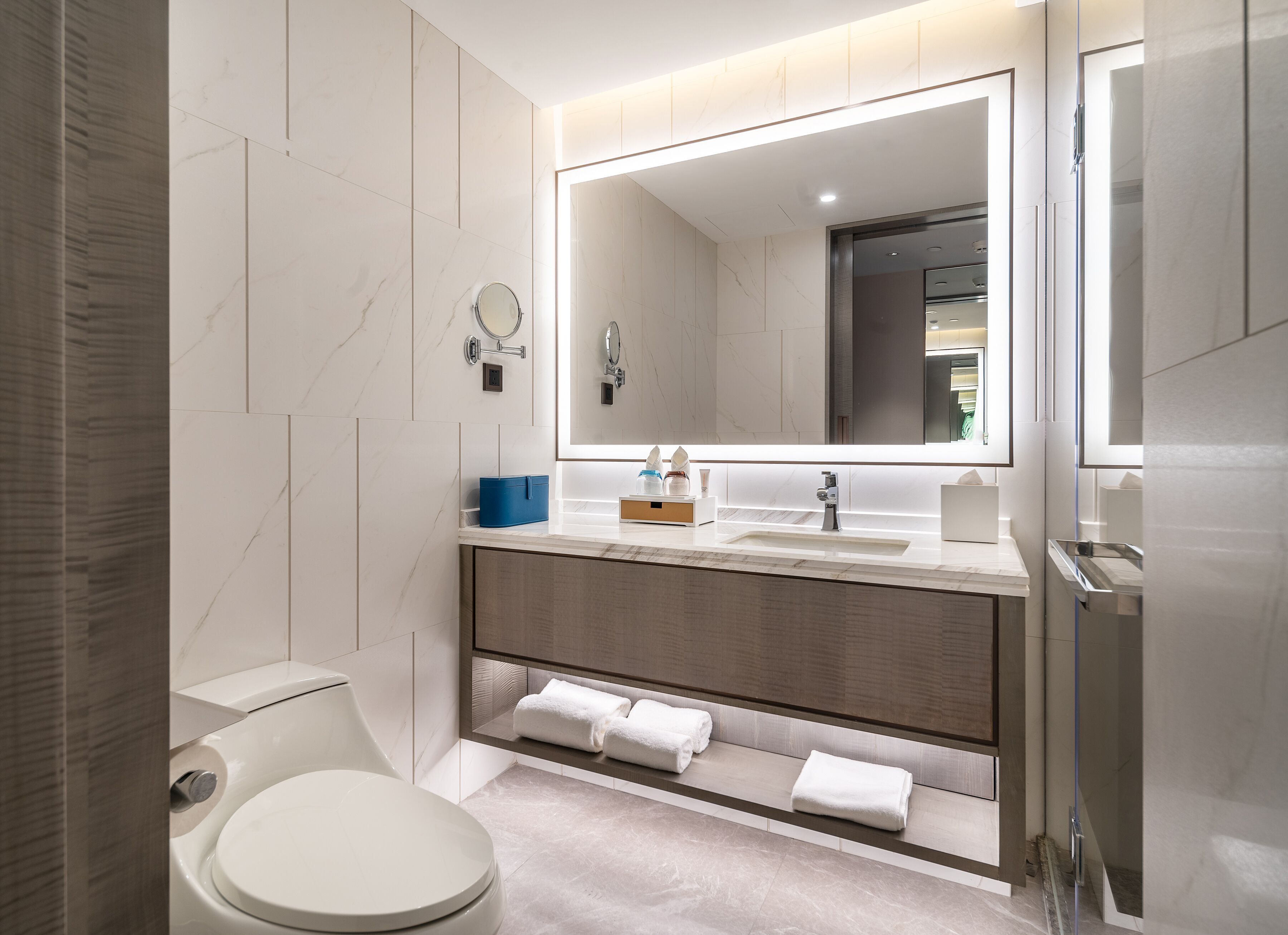 Un baño moderno con inodoro, lavamanos y espejo. Hay toallas y artículos de aseo en la encimera.