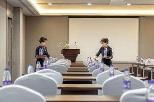 Un salón de conferencias con hileras de sillas blancas, un atril con un micrófono y botellas de agua sobre las mesas.