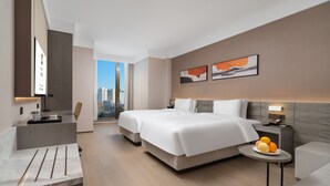 Habitación de hotel moderna con dos camas, una mesita con fruta, una mesita de noche y vista a la ciudad.