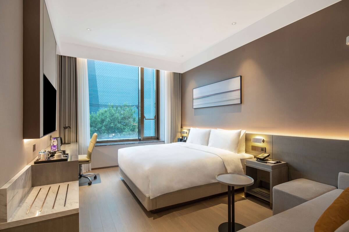 A modern hotel room with a large bed, bedside tables, a desk with a chair, a TV, and a window with a view of greenery.
