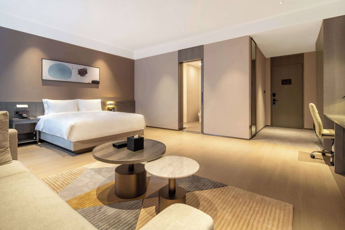 A modern hotel room with a large bed, a desk, a chair, and a small table.