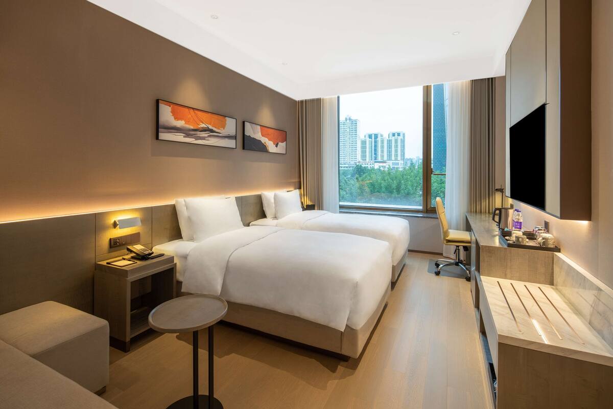 A modern hotel room with a large bed, bedside tables, a small round table, a TV, and a city view.
