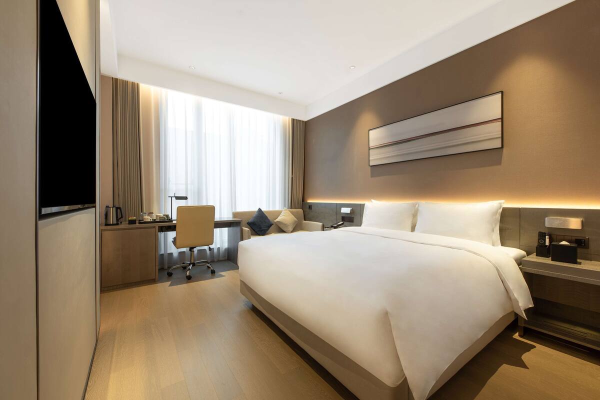 A modern hotel room with a large bed, a desk with a chair, a television, and a large window with curtains.