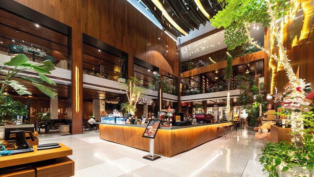 Un lobby amplio con un mostrador de recepción de madera, varias zonas de estar y plantas de interior.