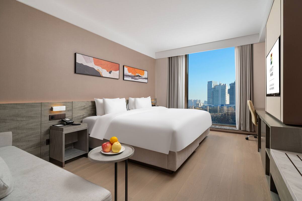 A modern hotel room with a large bed, a bedside table with fruit, a desk, and a city view.
