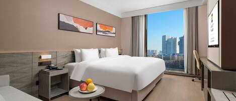 A modern hotel room with a large bed, a bedside table with fruit, a desk, and a city view.