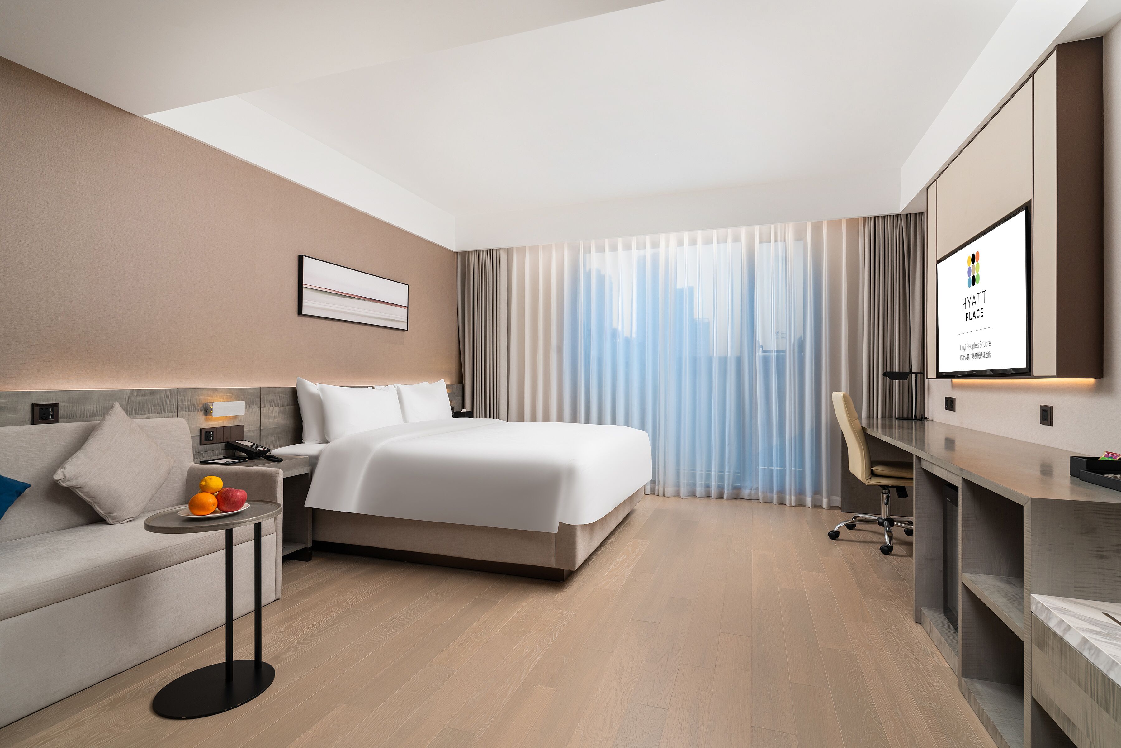 A modern hotel room with a bed, a desk, a chair, a small table with fruit, and a large window with curtains.