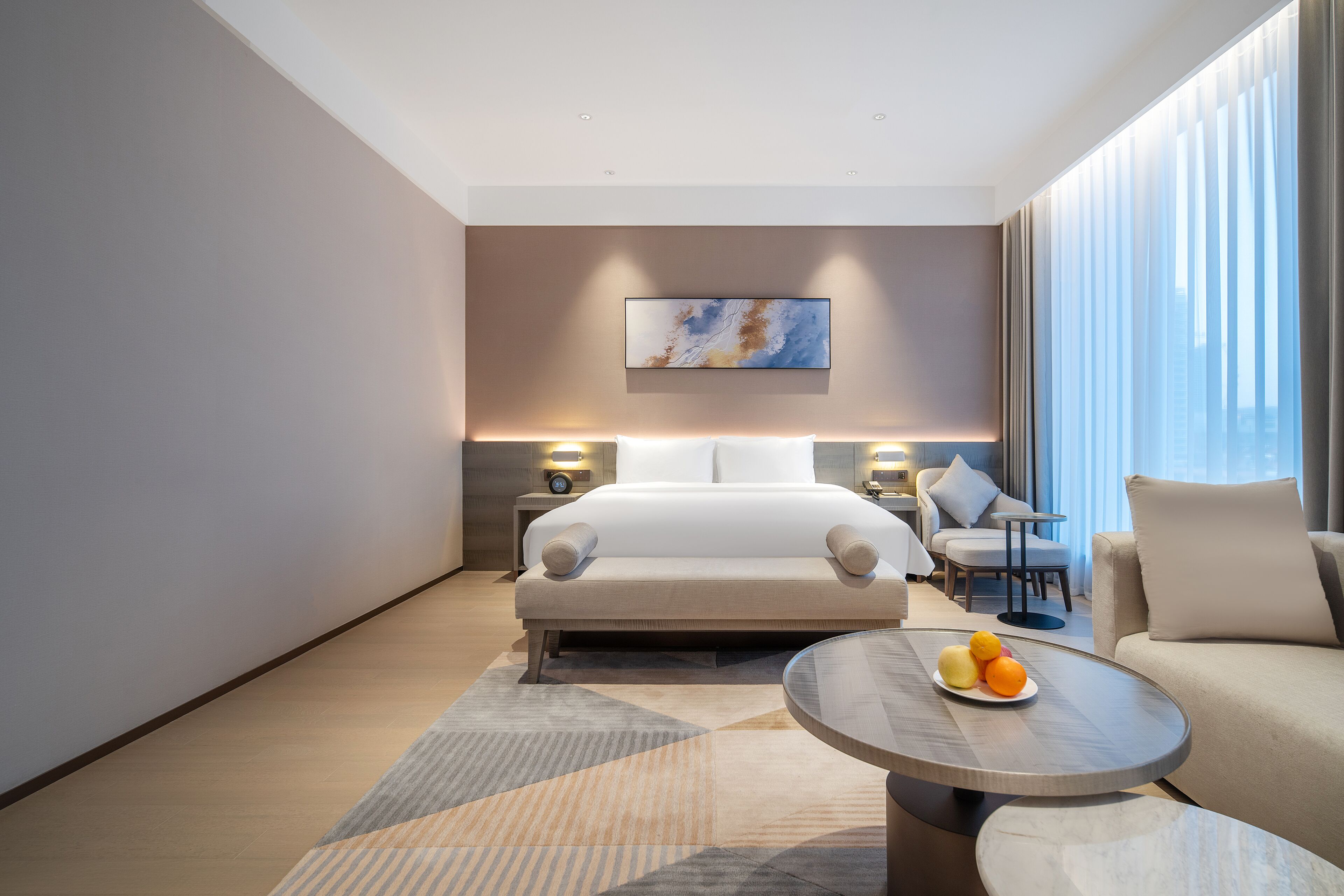 Habitación de hotel moderna con una cama grande, un sofá, una mesita de centro con fruta y vistas a la ciudad.