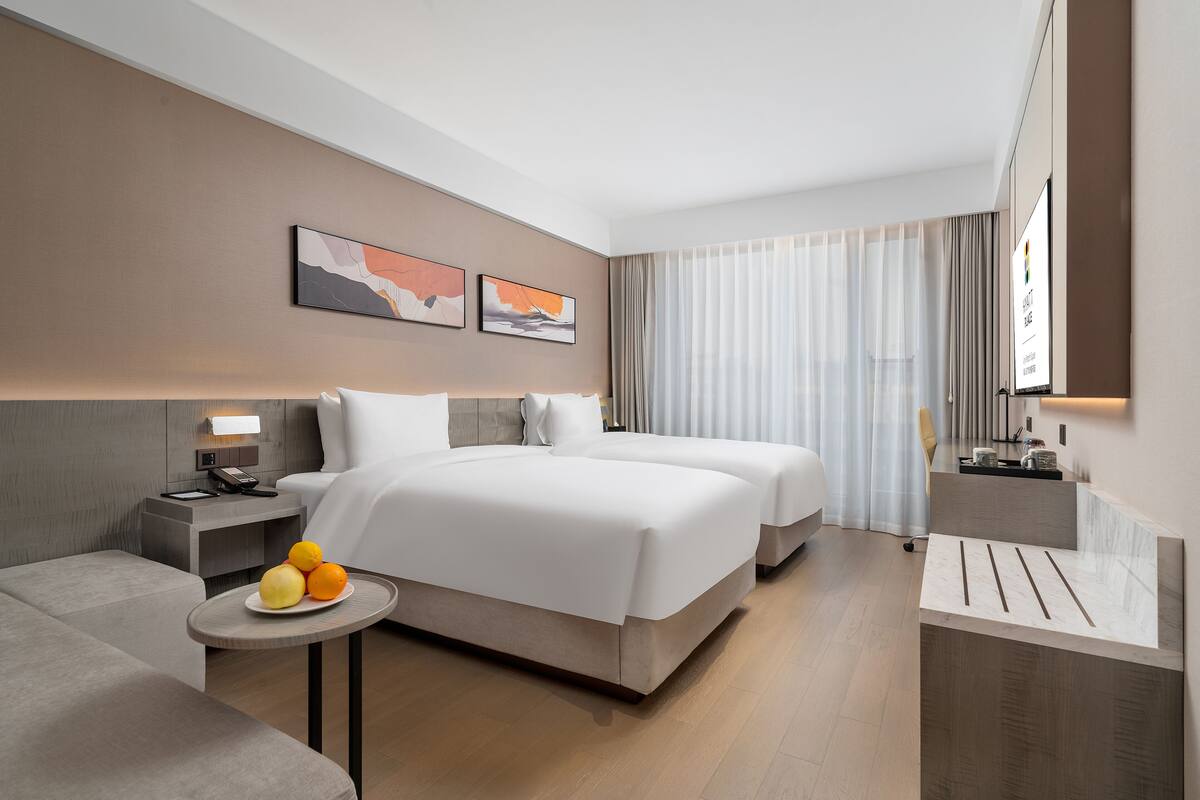 A modern hotel room with a large bed, bedside tables, a small round table with fruit, and a TV on the wall.