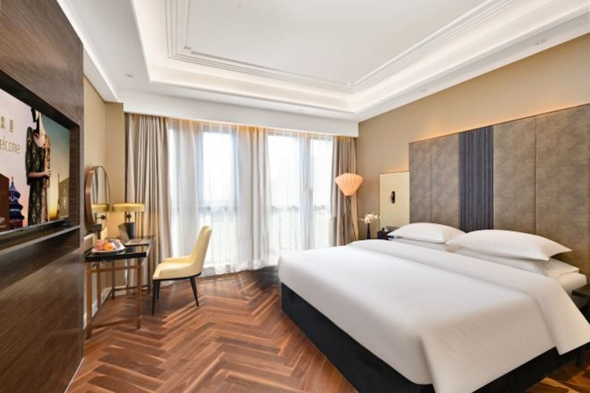 Habitación de hotel con una cama grande, un escritorio con silla, una lámpara y vistas al exterior a través de amplias ventanas.