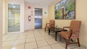 Un hall con suelo de baldosas, dos sillones a rayas, una mesita, un ascensor y un cuadro en la pared.
