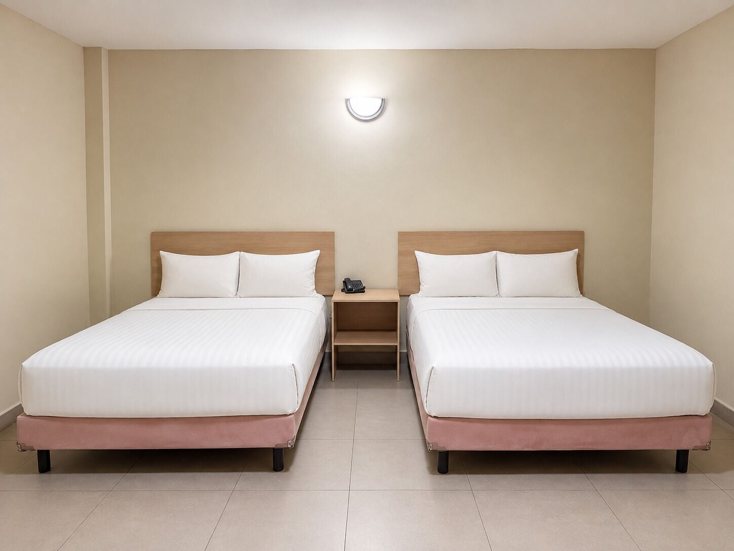 A hotel room with two beds, a bedside table with a phone, and a wall-mounted light fixture.