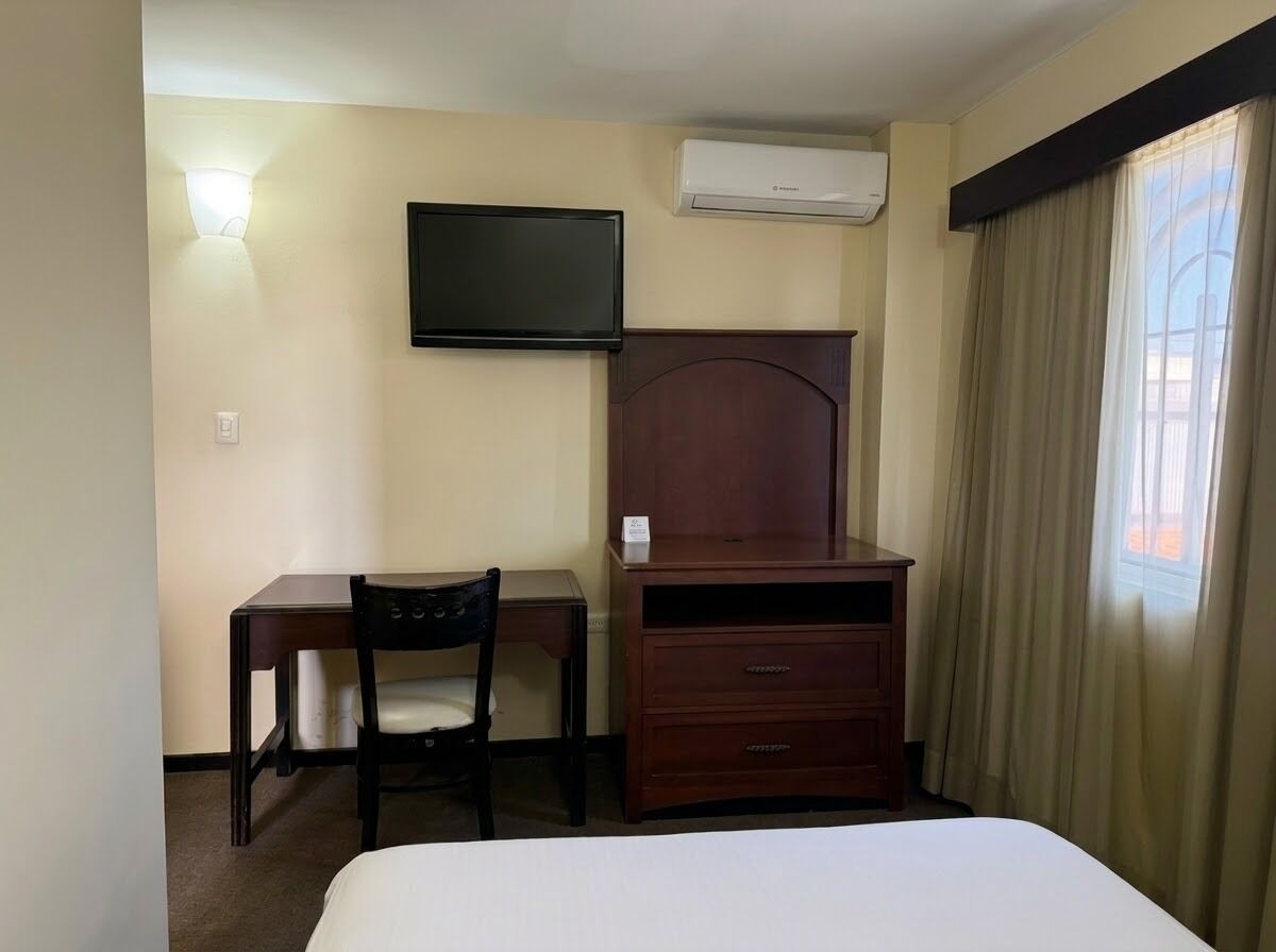 Habitación de hotel con cama, escritorio con silla, televisión, equipo de aire acondicionado y ventana con cortinas.