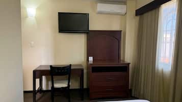 Habitación de hotel con cama, escritorio con silla, televisión, equipo de aire acondicionado y ventana con cortinas.