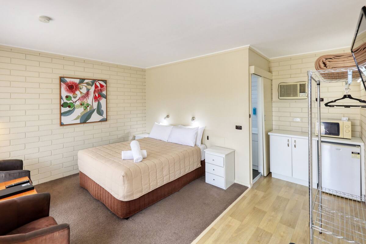 Una habitación de hotel con cama, mesitas de noche, un escritorio con silla, un cuadro en la pared y una kitchenette equipada con microondas y refrigerador.