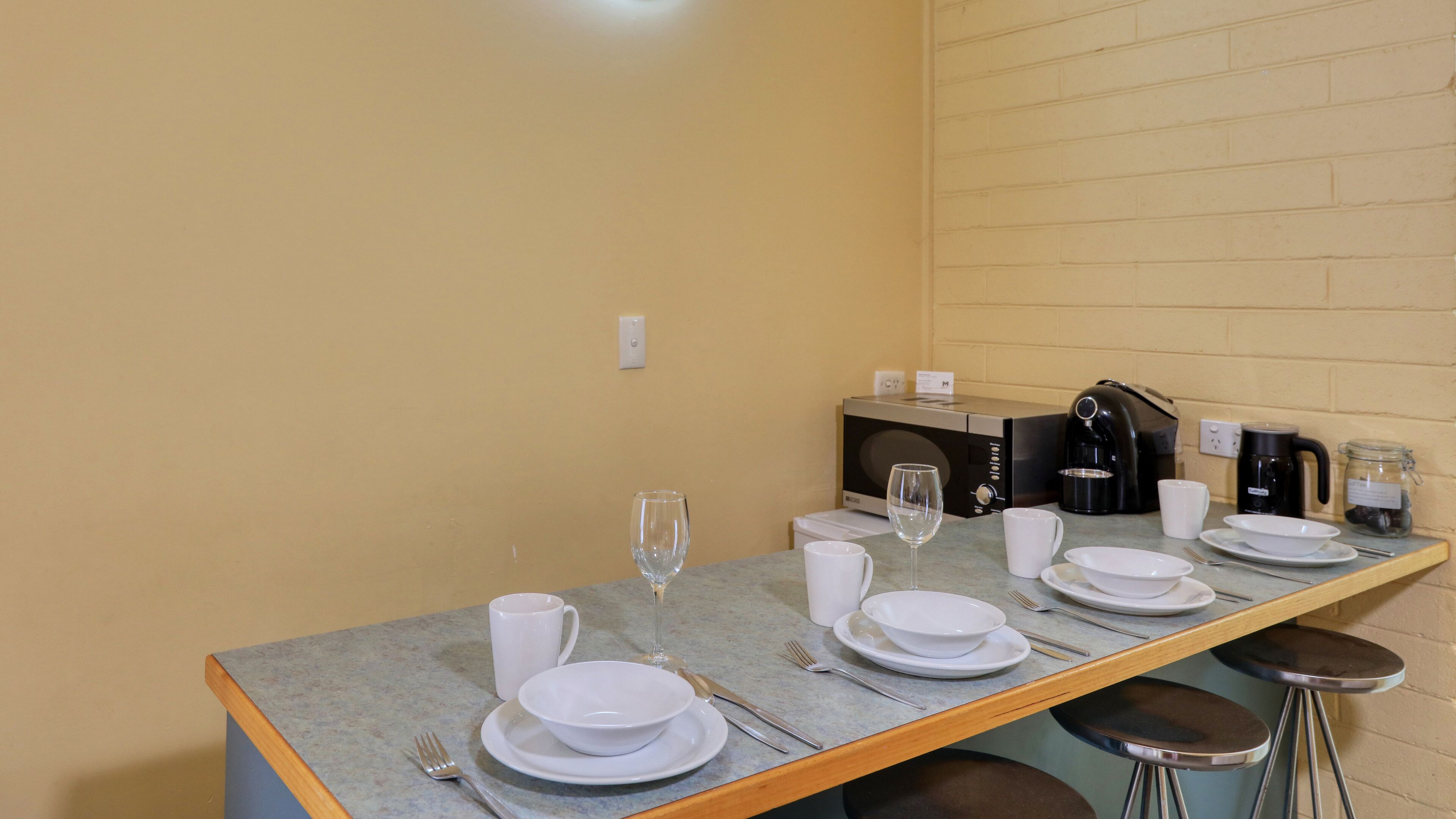 Comedor con una mesa dispuesta para cuatro, con platos, tazones, vasos y cubiertos. Hay una cafetera y un tarro de bayas sobre la mesa.