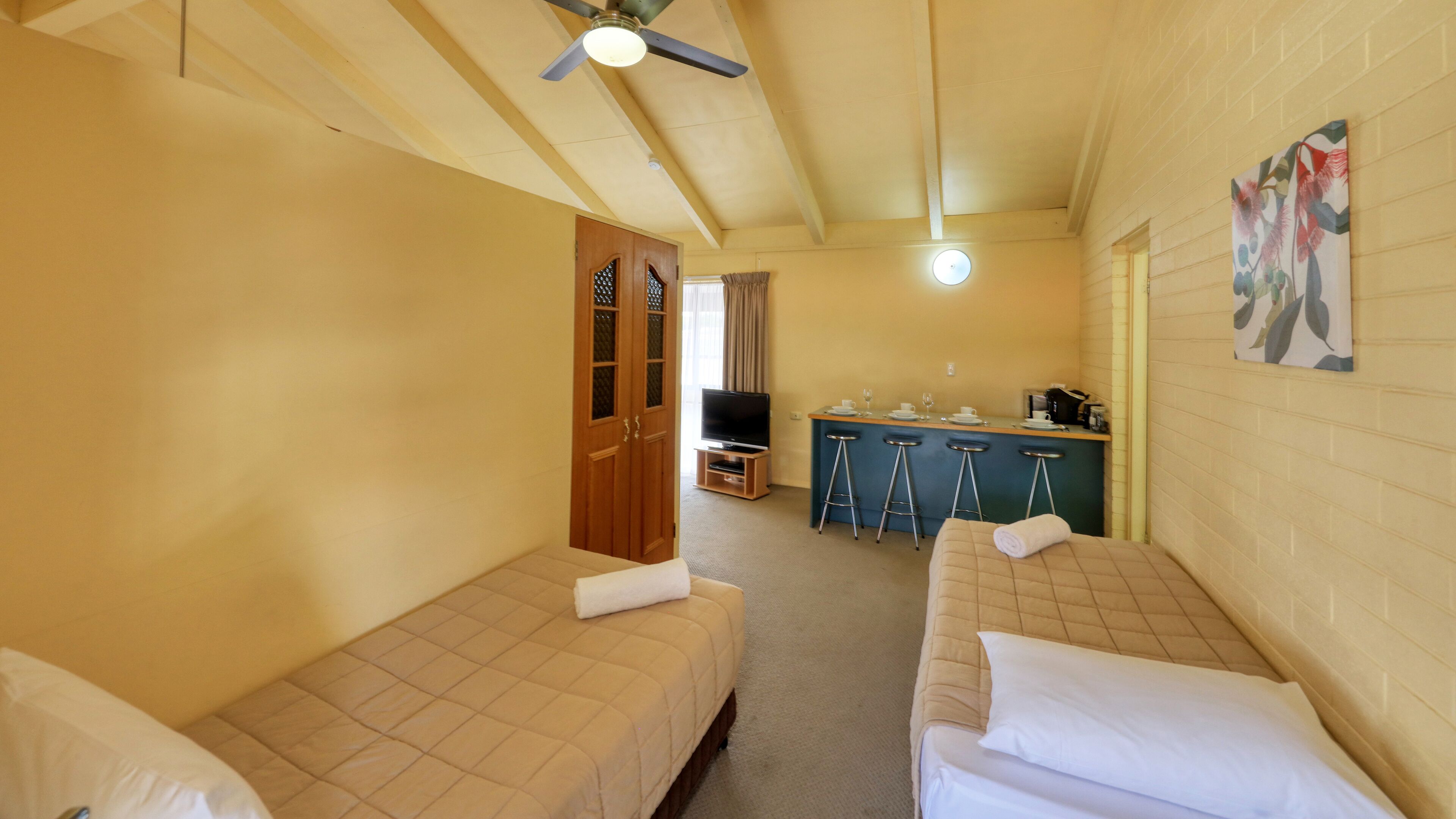 A compact hotel room with two single beds, a small kitchenette, a TV, and a ceiling fan.