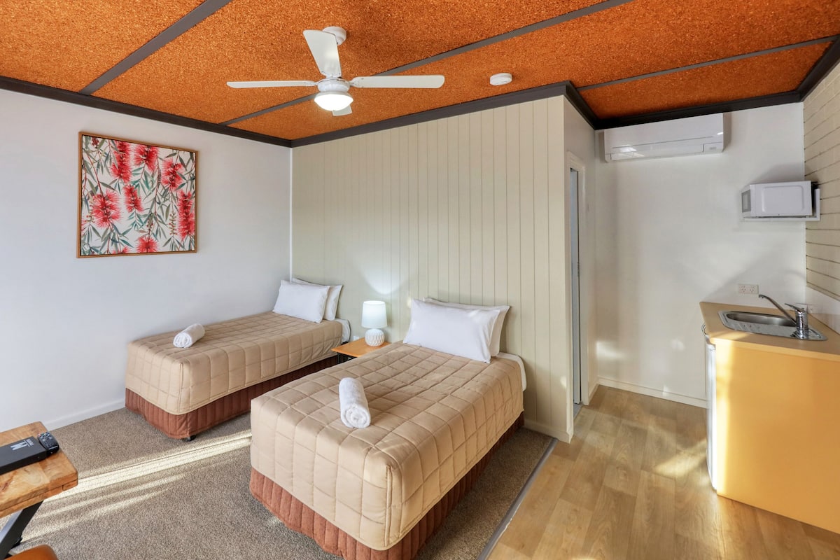 Habitación de hotel con dos camas, ventilador de techo, un cuadro, escritorio con control remoto y kitchenette.