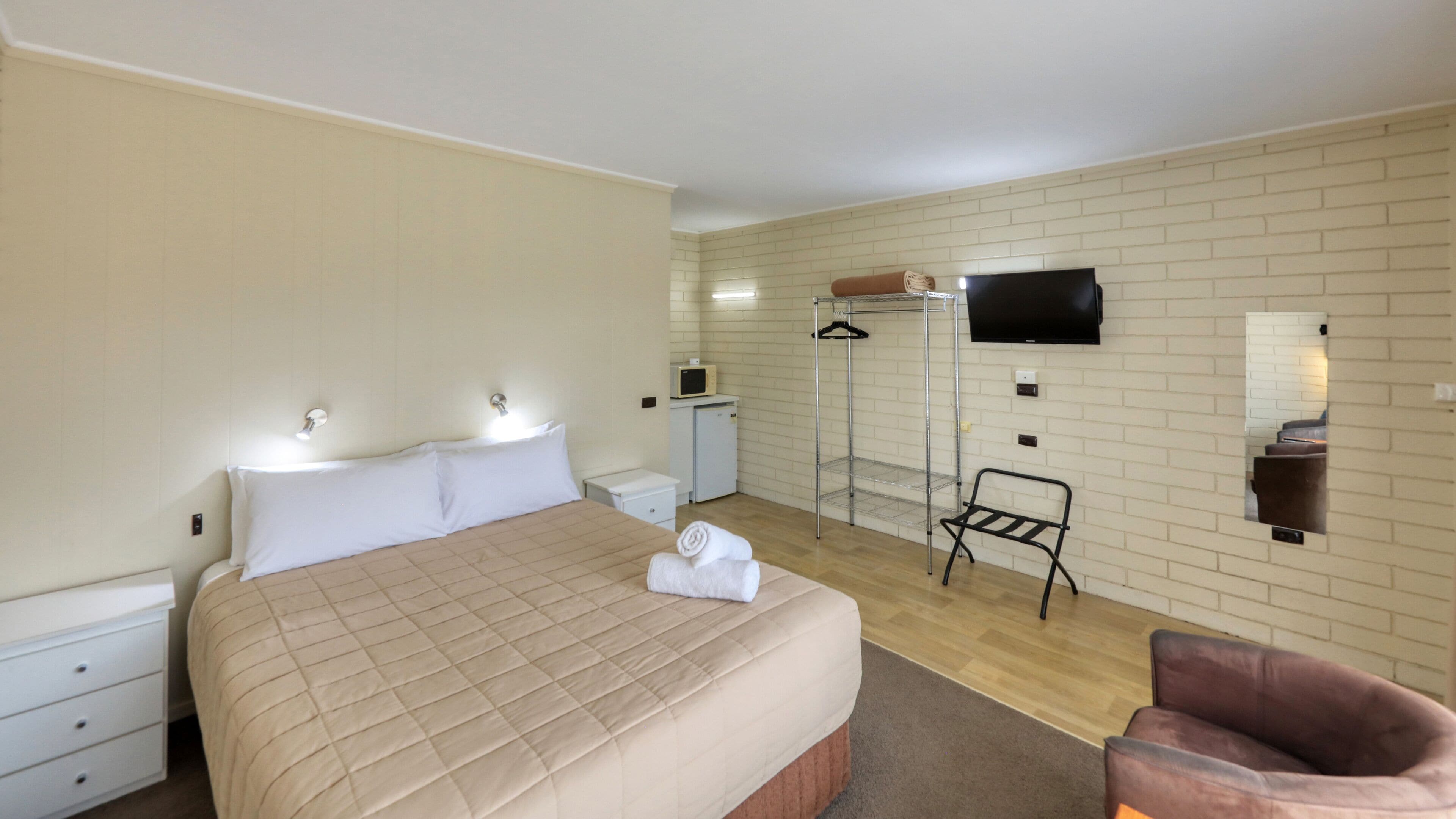 A hotel room with a bed, a chair, a TV, a mirror, a wardrobe, and a small desk with a lamp.