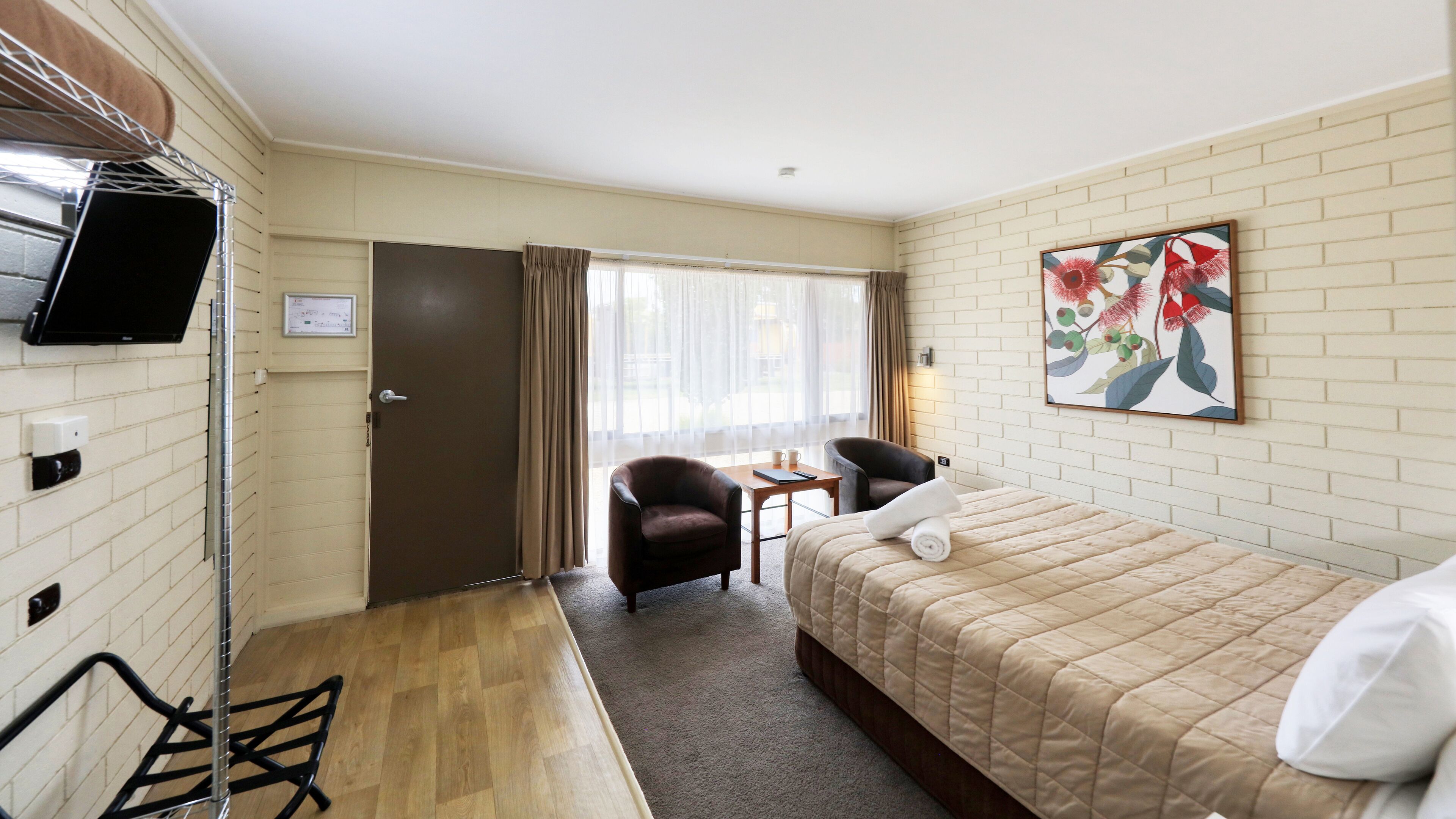 Quarto de hotel com cama, uma cadeira, uma mesinha, televisão e janela com cortinas.