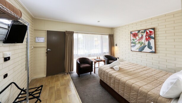 A hotel room with a bed, a chair, a small table, a TV, and a window with curtains. - Motel Meneres (Corowa)